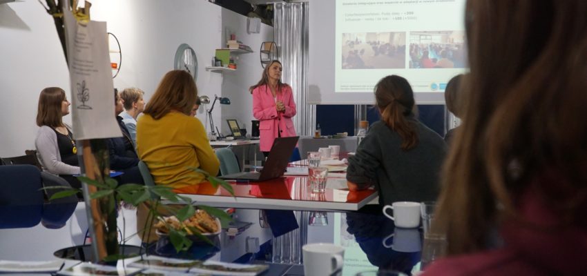 zdjęcie przedstawia scenę prezentacji na rzutniku - przedstawia kobieta w różowej marynarce, wokół stołu na pierwszym planie siedzą 3 osoby. W tle znajduje się ekran z wyświetlaną prezentacją.