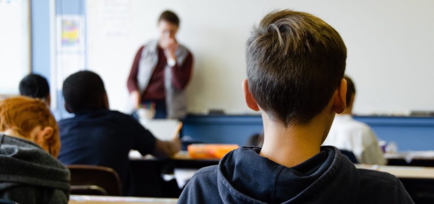 Zdjęcie przedstawia scenę z klasy skzoły podstawowej. W tle pod tablicą stoi nauczycielka. Na pierwszym i drugim planie widzimy sylwetki dwóch uczniów.